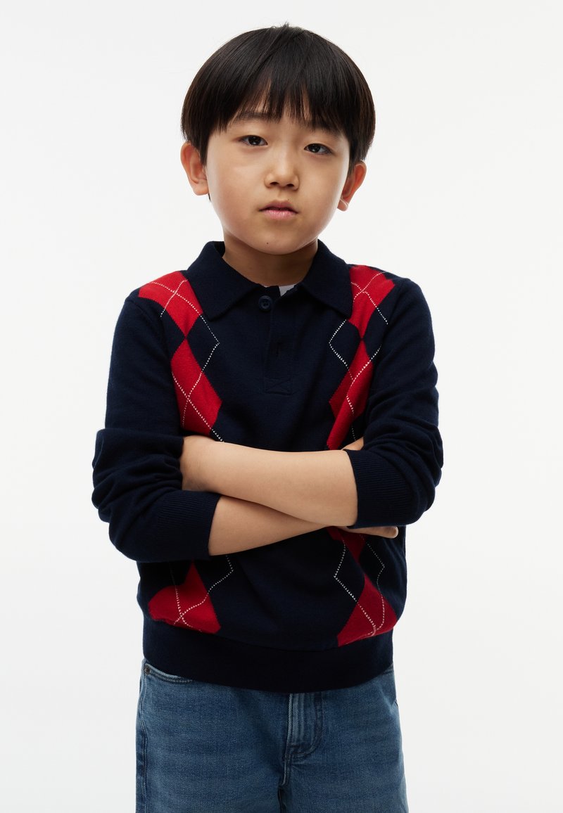 Jeune garçon aux cheveux noirs portant un pull bleu marine avec des motifs losanges rouges, bras croisés, debout devant un fond blanc uni.