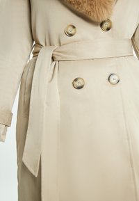 Trench coat beige con colletto in pelliccia, design doppiopetto, grandi bottoni rotondi e cintura in tessuto coordinato, caratterizzato da una texture liscia.