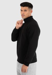 Zwart fleece trui met een hoge kraag, geribde manchetten en een gladde textuur. Model draagt grijze sweatpants. Minimale branding zichtbaar.
