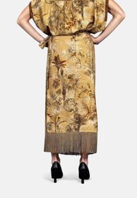 Abito in tessuto dorato con stampa tropicale, caratterizzato da maniche corte, vita legata e frange decorative all'orlo. Indossato con tacchi neri.