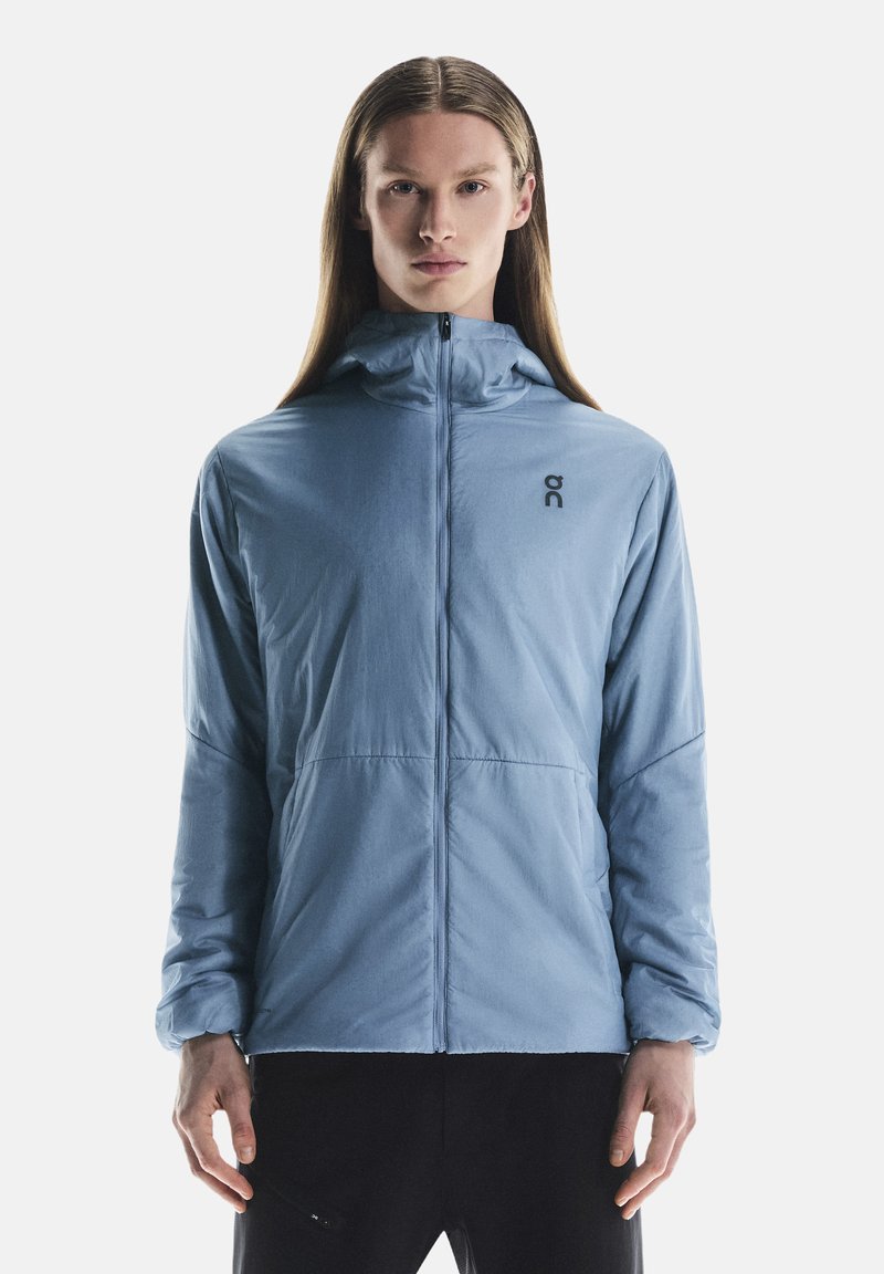 Veste zippée bleu clair avec capuche, fabriquée en tissu lisse. Présente un logo discret sur la poitrine et des poignets élastiques. Design simple et fonctionnel.