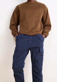 Brązowa bluzka z długim rękawem, dekoltem w kształcie litery V i raglanowymi rękawami, w połączeniu z granatowymi spodniami bojówkami z bocznymi kieszeniami. Gładka faktura.