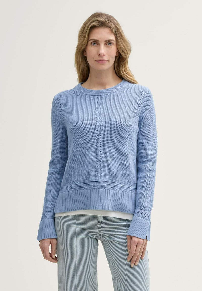 Hellblauer gestrickter Pullover mit rundem Ausschnitt, gerippten Ärmelbündchen und subtilen Texturmuster; über einem weißen Oberteil getragen und mit gestreiften Hosen kombiniert.