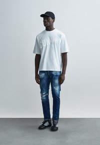 Λευκό κοντομάνικο μπλουζάκι με υφασμένο λογότυπο, συνδυασμένο με τζιν παντελόνια σε μπλε ξεβαμμένο χρώμα και μαύρα παπούτσια με λευκές ρίγες· φορεμένο μαύρο καπέλο.