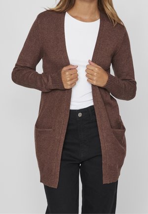 Femme portant un cardigan marron à ouverture frontale avec poches, sur une chemise blanche et un pantalon noir, sur un fond uni.