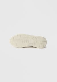 Sole de teniși din cauciuc cu un model texturat, având o culoare crem deschis și vârf rotunjit. Design al benzii de rulare în formă de grilă pentru aderență.