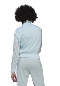 Persona con cabello rizado vistiendo un chándal azul claro, de espaldas, con las manos relajadas a los lados contra un fondo blanco.