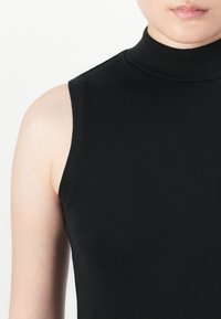 Kratki prikaz osobe koja nosi crni turtleneck bez rukava, prikazujući vrat, rame i djelomično lice.