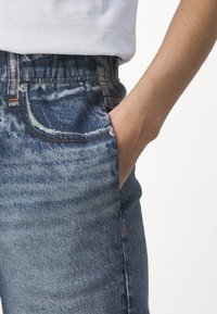 Blauwe denim jeans met een versleten textuur, voorzien van een elastische tailleband, voorzakken en contrasterende oranje stiksels.