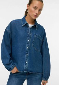 Giubbotto di jeans in blu medio con chiusura frontale a bottoni, tasca a toppa, vestibilità oversize e maniche lunghe. Tessuto testurizzato con dettagli di cucitura sottili.