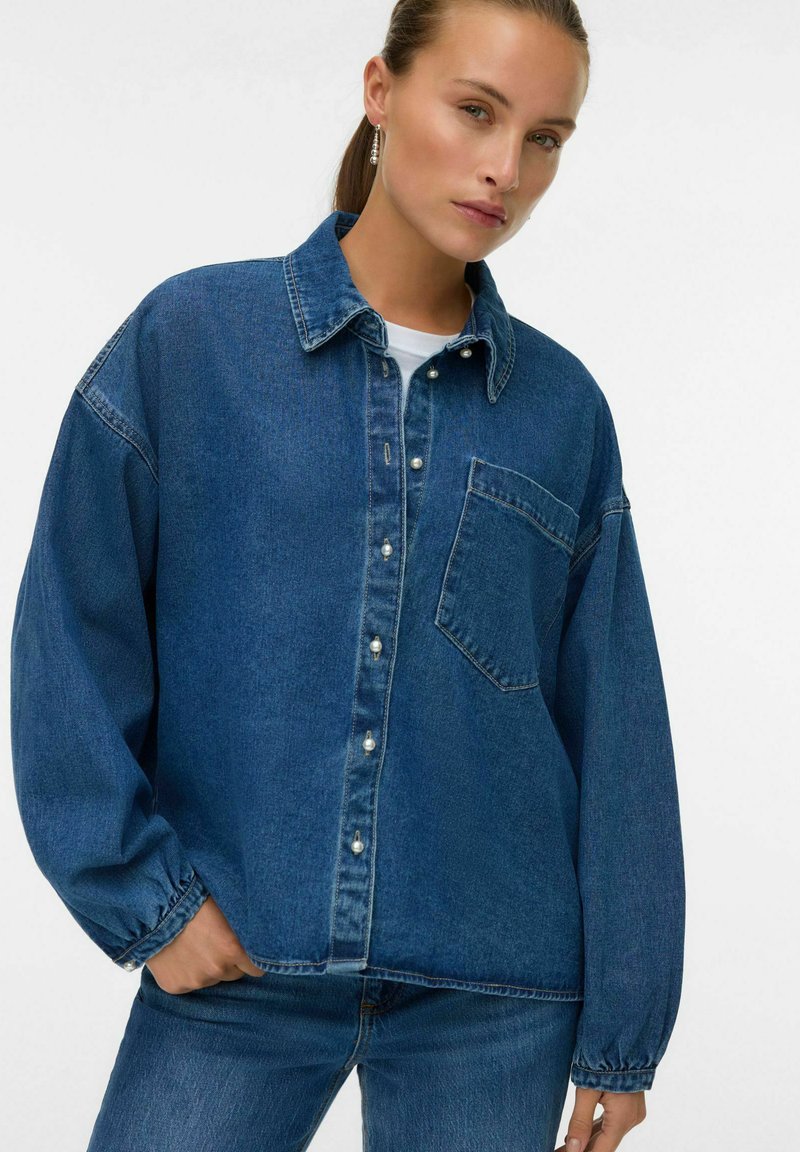 Denim jakna v srednje modri barvi z gumbastim zapiranjem spredaj, nastavljivim žepom, širokim krojem in dolgimi rokavi. Teksturovana tkanina z diskretnimi šivnimi detajli.