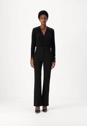 Tuta nera con design avvolgente, maniche lunghe con dettagli a bottone e pantaloni a gamba larga sartoriali. Texture liscia e vita aderente.