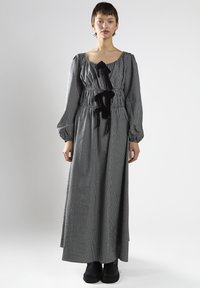 Mujer vestida con un largo vestido a cuadros en blanco y negro con mangas abullonadas y lazos negros, combinado con botas negras, de pie frente a un fondo blanco.