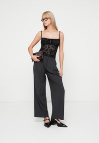 Top in pizzo floreale nero con spalline sottili, abbinato a pantaloni grigi a vita alta e gamba larga e scarpe nere con punta affilata, mentre tiene una pochette.