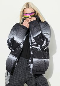 Chaqueta de plumas en blanco y negro con un patrón de puntos, que presenta un cierre de cremallera, cuello alto y puños elásticos. Combinada con pantalones negros.