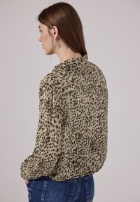 Woman with brown hair wearing silver hoop earrings, a beige and black leopard-print blouse, and blue jeans, facing sideways with hands in pockets.
