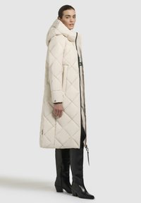 Lunga giacca trapuntata di colore beige chiaro con cappuccio. Caratterizzata da cuciture diagonali, tasche laterali e una chiusura con zip. Abbinata a stivali neri alti fino al ginocchio.