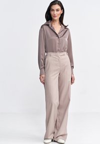 Koszulka w kolorze taupe o jedwabistej fakturze z kołnierzykiem i zapięciem na guziki, zestawiona z jasnymi beżowymi spodniami w szerokie paski. Buty w kolorze off-white.