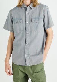 Camicia in denim a maniche corte grigio chiaro con chiusura a bottoni a pressione, due tasche sul petto e orlo dritto. Texture morbida con una leggera sgualcitura.