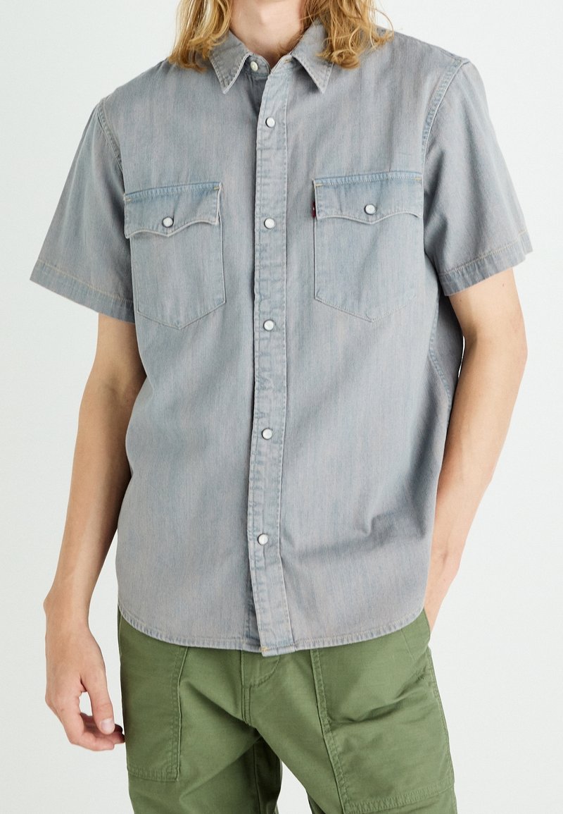 Camicia in denim a maniche corte grigio chiaro con chiusura a bottoni a pressione, due tasche sul petto e orlo dritto. Texture morbida con una leggera sgualcitura.