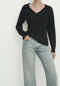 Personne portant une chemise noire à manches longues avec col en V et un jean bleu clair taille haute, debout avec une main dans la poche.