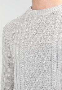 Ljusgrå stickad tröja med flät- och ribbmönster, som har en rund halsringning, på en persons överkropp.