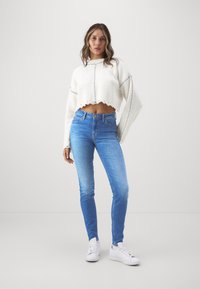 Vit kortärmad tröja med ribbad textur, kombinerad med ljusblå skinny jeans. Modellen har vita sneakers på sig, vilket ger en avslappnad outfit.