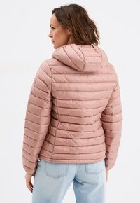 Giacca a vento imbottita rosa chiaro con cappuccio, caratterizzata da cuciture a righe orizzontali. Presenta una forma affusolata e una consistenza morbida e lucida. Abbinata a jeans blu.