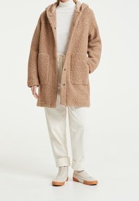 Abrigo de forro polar marrón con capucha, superficie texturizada, grandes bolsillos frontales y botones a presión, combinado con pantalones de pana color crema y zapatos de caña alta beige.