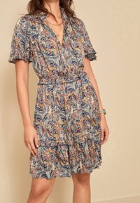 Femme portant une robe à manches courtes et décolletée en V, avec un motif cachemire multicolore et un ourlet volanté, accessoirisée de colliers superposés et d'une montre-bracelet.