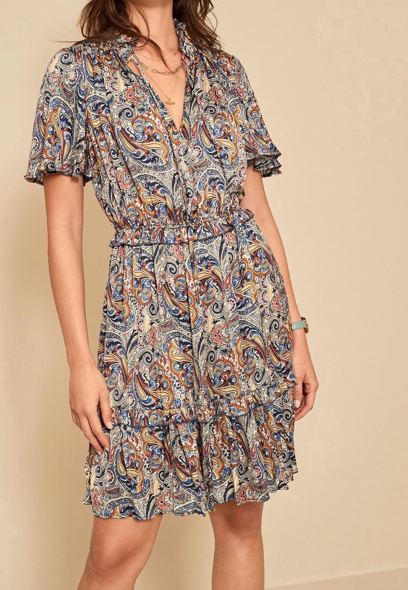 Femme portant une robe à manches courtes et décolletée en V, avec un motif cachemire multicolore et un ourlet volanté, accessoirisée de colliers superposés et d'une montre-bracelet.