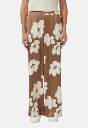 Bruine broek met wijde pijpen en grote witte en lichtroze bloemenprints, gedragen met witte schoenen en een crème top in de broek gestopt.