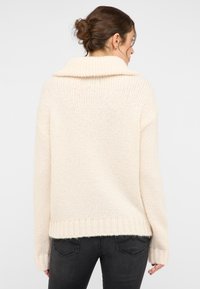 Przesadnie duży kremowy sweter dzianinowy z zawiniętym kołnierzem, ściągaczami przy mankietach i u dołu. Posiada teksturowany wzór ściegu oraz luźny krój.