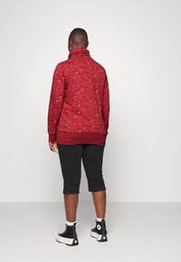 Person iført en rød jakke med fuglemotiv, sorte knælange bukser, hvide sokker og sorte høj-top sneakers, stående med ryggen mod kameraet.