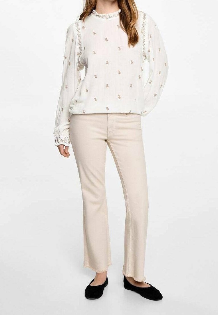 Femme portant un chemisier blanc à manches longues avec de petits motifs floraux, un pantalon beige court aux ourlets effilochés, et des chaussures plates noires.