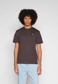 Brun crew neck t-skjorte laget av bomull, med en liten grønn logo på venstre bryst, korte ermer og en avslappet passform.