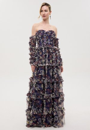 Vrouw draagt een lange strapless jurk met ruches en bloemenprint, gemaakt van doorschijnende, gestructureerde stof, staand tegen een effen witte achtergrond.