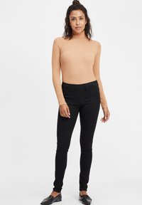 Bordo turtleneck majica u breskvastoj boji, prilagođenog kroja. U paru s crnim uskim hlačama i crnim mokasinkama, stoji na jednoličnoj pozadini.