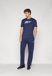 Sötétkék pamut póló fehér Reebok logóval, hozzáillő sötétkék melegítőnadrággal és fekete sportcipővel párosítva. Egyszerű dizájn, laza szabás.