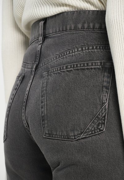 Primer plano de una persona que lleva unos jeans de mezclilla negros desgastados de cintura alta y un suéter de canalé color crema.