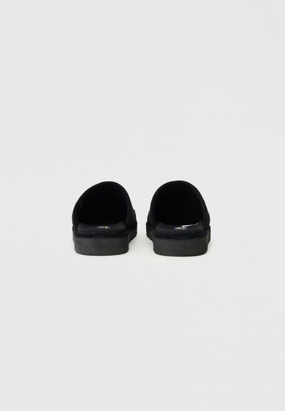 Μαύρες παντόφλες με μαλακό σουέντ άνω μέρος, ανοιχτή σχεδίαση πίσω και επίπεδες ριγωτές σόλες. Ελάχιστες λεπτομέρειες και υφή.