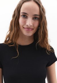 Černé žebrované tričko s krátkým rukávem a kulatým výstřihem; hladká textura a těsný střih. Modelka má vlnité vlasy a minimální líčení.