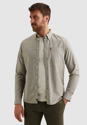 Mężczyzna z brodą, ubrany w rozpiętą szarą koszulę na białej koszulce i ciemnozielonych spodniach, patrzący w lewo, z jedną ręką w kieszeni.