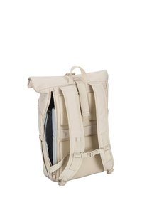 Sac à dos beige à rabat avec des bretelles rembourrées, un panneau arrière en maille, une boucle de poitrine et une poche latérale partiellement zippée montrant des objets gris et noirs à l'intérieur.