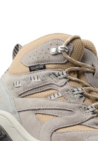 Scarpa da trekking beige e grigia con tomaia testurizzata, accenti in rete e una punta arrotondata prominente. Presenta un design con lacci e occhielli rinforzati.