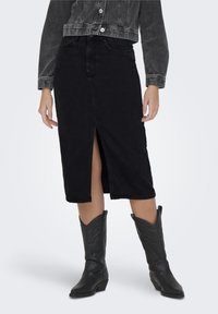 Jupe en denim noir avec fente frontale, longueur genoux. Associée à des bottes de cow-boy noires avec des détails gaufrés. Style élégant avec un minimum de détails.