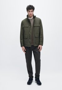 Olivengrøn jakke med høj krave, to brystlommer og knaplukning, kombineret med mørke bukser og sorte sneakers.