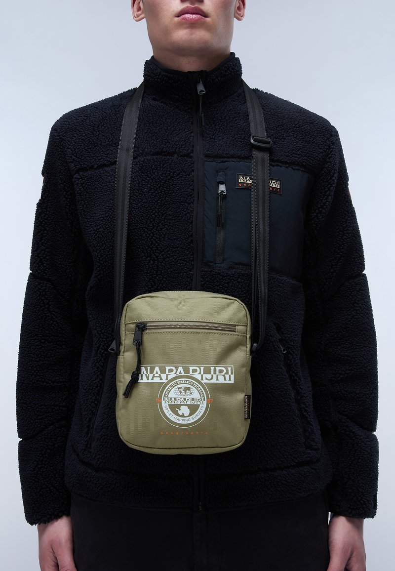Borsa a tracolla verde oliva con chiusura a zip, caratterizzata da logo stampato bianco e design circolare. Indossata sopra un giubbotto di pile nero.