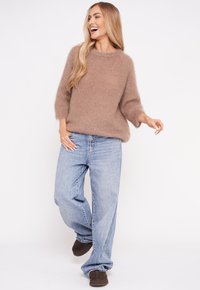 Eine Frau lächelt und geht, trägt einen flauschigen braunen Pullover, lockere hellblaue Jeans und dunkelbraune Schuhe vor einem weißen Hintergrund.