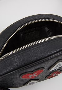 Svart strukturerat läderväska med dragkedja; insidan har en slät foder. Framsidan visar färgglada glittrande hjärtan och diamantmotiv.